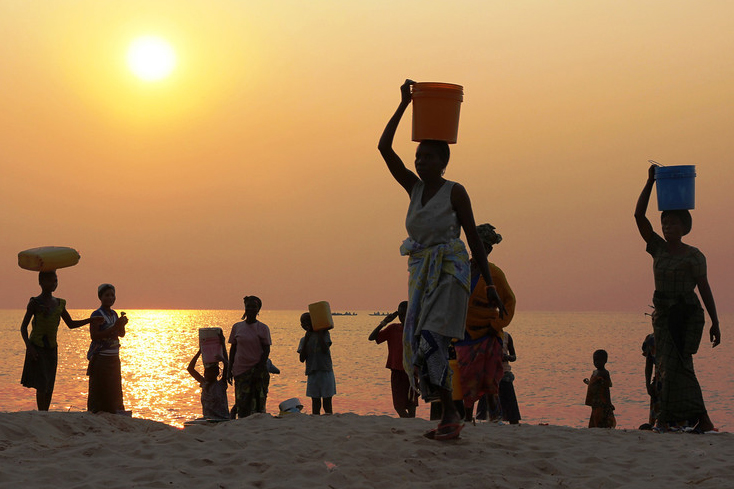 Over a dozen women carry water from Tanganyika Lake as the sun rises in the background.
