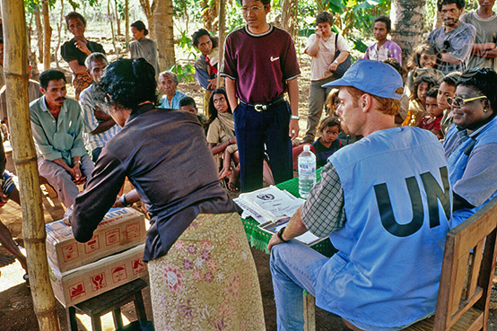 Voluntarios de la ONU en Timor Oriental en una mesa con un gran grupo de timorenses orientales al fondo.